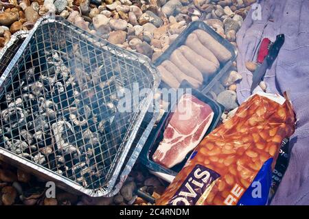 Traditionelle englische Würstchen und Speck zum Frühstück, das auf einem Einweg-Grill am Strand von Normans Bay, West Sussex zubereitet wird Stockfoto