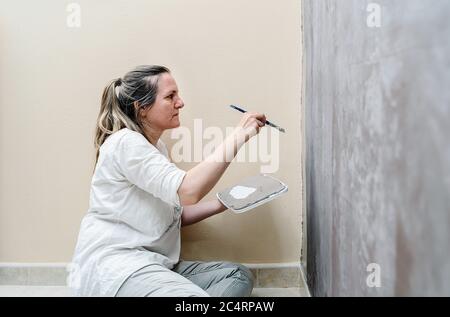 Blonde barfuß Frau sitzt auf ihrem Boden beginnt malen eine braune Wand in eine graue Farbe mit einem kleinen Pinsel. Die Frau trägt ein weißes Hemd und ein Stockfoto