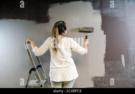 Frau kletterte eine Leiter und nahm eine Malrolle voll grauer Malerei an einer braunen Wand. Maler ist auf und ab Walze decken die Wand mit Stockfoto