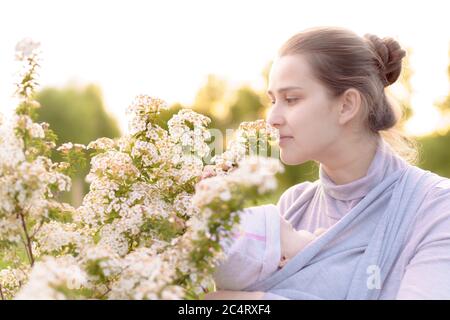 Mutterschaft, Pflege, Säuglinge, Sommer, Elternkonzept - Junge schöne Mutter mit einem Neugeborenen in einem Tragetuch geht und schaut auf ein Kind im Hintergrund Stockfoto