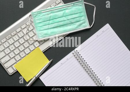 Schreibtisch mit Computer-Tastatur, Notizblock, Stift, leeres gelbes Papier und medizinischer Gesichtsmaske. Draufsicht oder flach liegend. Stockfoto