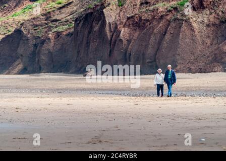 Ein älteres Paar geht über einen leeren Strand am Fuß von hohen Klippen und hält die Hände. Stockfoto