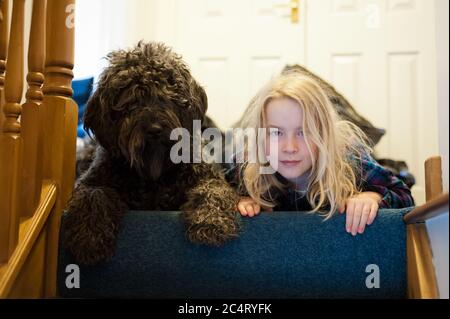 Ein junges blondes Mädchen und ein zackiger schwarzer Labradoodle, der von der Treppe nach unten schaut. Stockfoto