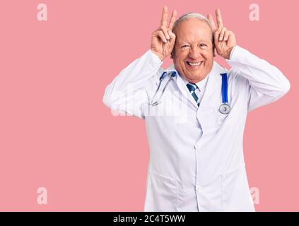 Senior stattlich grauhaarige Mann trägt Doktor Mantel und Stethoskop posiert lustig und verrückt mit Fingern auf dem Kopf als Hase Ohren, lächelnd fröhlich Stockfoto