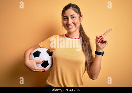Schöne Brünette Spieler Frau Fußball spielen mit Fußball auf gelbem Hintergrund sehr glücklich zeigt mit Hand und Finger zur Seite Stockfoto