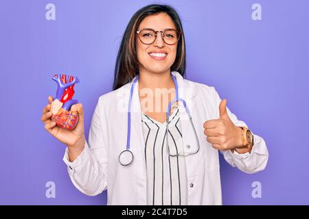 Junge Arzt Kardiologie-Fachfrau hält medizinisches Herz über prüple Hintergrund glücklich mit großem Lächeln tun ok Zeichen, Daumen mit Fingern, Excel Stockfoto
