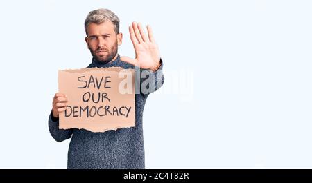 Junge blonde Mann halten speichern unsere Demokratie Karton Banner mit offener Hand tun Stop-Zeichen mit ernsten und selbstbewussten Ausdruck, Verteidigung Geste Stockfoto