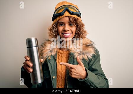 Junge afroamerikanische Skifahrerin Frau trägt Skibrille trinken Thermo mit Kaffee sehr glücklich zeigen mit Hand und Finger Stockfoto