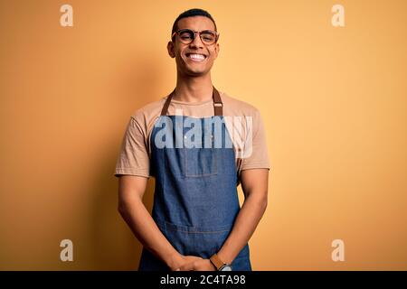 Junger hübscher afroamerikanischer Ladenbesitzer mit Schürze auf gelbem Hintergrund und einem fröhlichen und coolen Lächeln im Gesicht. Glücklicher Mensch. Stockfoto