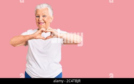 Ältere schöne Frau mit blauen Augen und grauen Haaren tragen lässige weiße T-Shirt lächelnd in der Liebe tun Herz Symbol Form mit Händen. Romantisches Konzept. Stockfoto