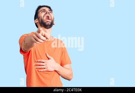 Junger, hübscher Mann mit Bart, der ein lässiges T-Shirt trägt, lacht über dich und zeigt mit dem Finger auf die Kamera, mit der Hand über den Körper, Schamgefühl Stockfoto