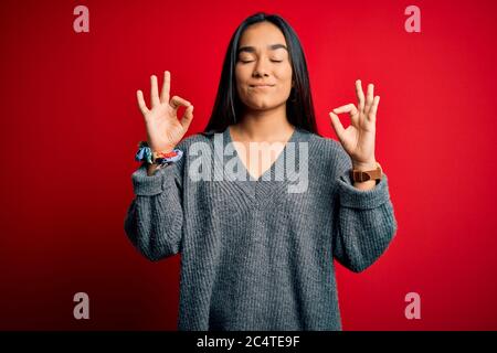 Junge schöne asiatische Frau mit legeren Pullover über isoliertem rotem Hintergrund entspannen Sie sich und lächeln Sie mit geschlossenen Augen, die Meditationsgeste w ausführen Stockfoto