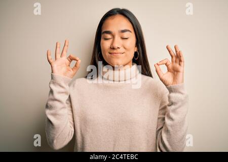 Junge schöne asiatische Frau trägt lässig Rollkragenpullover über weißem Hintergrund entspannen und lächeln mit geschlossenen Augen tun Meditation Geste mit f Stockfoto