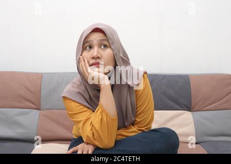 Schöne asiatische muslimische Frau denken etwas ernst, während auf dem Sofa sitzen, Mädchen mit Kontemplation Ausdruck, Blick nach oben Stockfoto