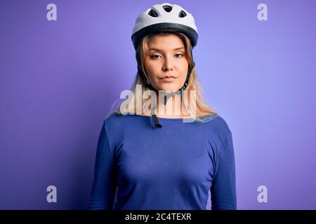 Junge schöne blonde Radfahrer Frau trägt Fahrrad Sicherheitshelm über lila Hintergrund mit ernsten Ausdruck auf Gesicht. Einfach und natürlich aussehende a Stockfoto