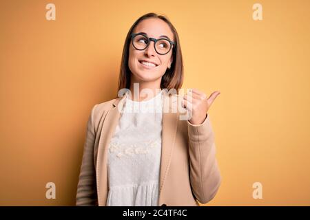 Junge schöne Geschäftsfrau tragen Jacke und Brille über isolierten gelben Hintergrund lächelnd mit glücklichen Gesicht suchen und zeigen auf die Seite mit Stockfoto