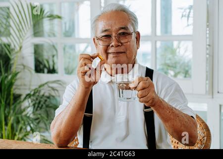 Senior Asian Mann mit Tasse Wasser und Pille Blick auf Kamera und nehmen Heilmittel in gemütlichen Raum zu Hause. Alter Mann nimmt Medizin. Stockfoto
