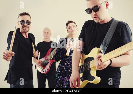 Stilvoller junger Mann in Sonnenbrille, der Bassgitarre spielt, seine Band spielt im Hintergrund Stockfoto