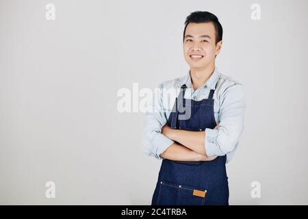 Porträt von gut lächelnden jungen asiatischen Kellner in Jeanschürze Falten Arme und Blick auf Kamera, isoliert auf grau Stockfoto