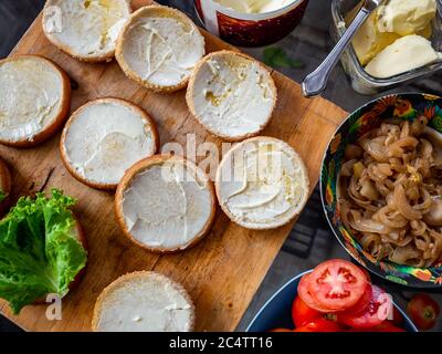 Prozess der Herstellung von leckeren hausgemachten Burger. Draufsicht auf Brötchen mit Butter, karamellisierte Zwiebeln, frische Tomaten auf Holzschneidebrett, vorbereitet zu machen Stockfoto