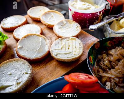 Prozess der Herstellung von leckeren hausgemachten Burger. Brötchen mit Butter, karamellisierte Zwiebeln, frische Tomaten auf Holzschneidebrett, zubereitet für hausgemachte BU Stockfoto