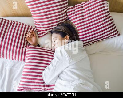Schöne glückliche asiatische Frauen kurze Haare tragen weißes Hemd schlafen mit rot weiß gestreiften Kissen auf dem Bett im Schlafzimmer im Haus am Morgen. Stockfoto