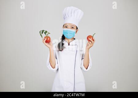 Positive junge weibliche Restaurant-Chef in medizinische Maske hält frische Tomaten für Sauce Stockfoto