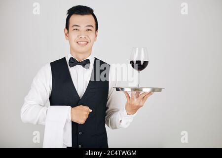 Positiver junger Kellner mit weißem Handtuch und einem Glas Rotwein auf Metalltablett Stockfoto