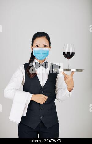 Junge seriöse Kellnerin trägt weißes Handtuch und Tablett mit Glas Wein, isoliert auf grau Stockfoto