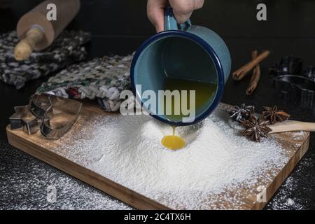 Eine Person gießt das Eigelb auf das Mehl auf einem Holztisch mit Zimtsternen und Backformen an den Seiten Stockfoto