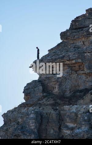 Ein Mann steht am Rande eines Berges vor einem blauen Himmel. Ein Tourist in den Bergen steht auf den Felsen und schaut hinunter. Abenteuer, Reisen, aktiver Lebensstil. Stockfoto