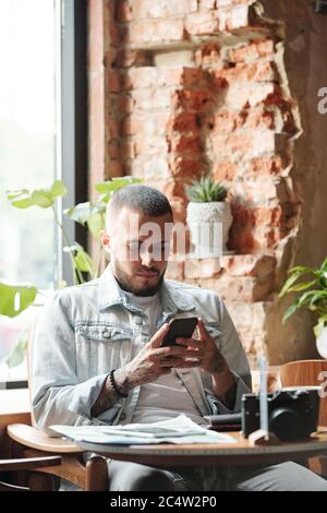 Serious junger Mann in Denim Jacke sitzen am Tisch im Loft-Café und mit Smartphone während SMS Stockfoto
