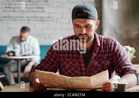 Konzentrierter junger bärtiger Mann in einer Ballkappe, der am Tisch sitzt und im Café Papierkarte verwendet Stockfoto