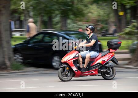 Belgrad, Serbien - 25. Juni 2020: Junger Mann in Shorts und T-Shirt auf rotem Roller mit hinterer Box auf der Stadtstraße, Seitenansicht Stockfoto