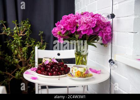 Bouquet von Pfingstrosen, zwei Tassen Tee und frische Kirschen auf weißem Tisch. Moderne Balkonausstattung. Stockfoto