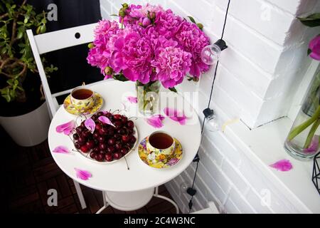 Bouquet von Pfingstrosen, zwei Tassen Tee und frische Kirschen auf weißem Tisch. Moderne Balkonausstattung. Stockfoto