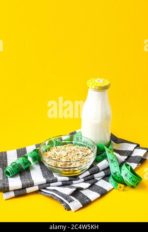 Haferflocken in einer Schüssel, Milch in einer Glasflasche und grünes Zentimeter-Band auf hellgelbem Hintergrund mit Küchentuch, Kopierraum Stockfoto