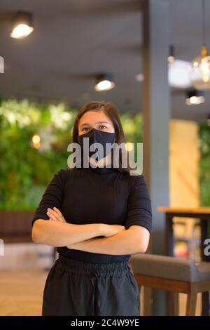 Junge asiatische Geschäftsfrau mit gekreuzten Armen in Maske und sozialer Distanzierung im Café Stockfoto