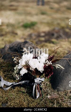 brautstrauß aus weißen Rosen, Lilie, Burgunder Nelken, protea, getrockneten Zweigen, Brunia und Leukadendron mit grauen und schwarzen Bändern auf dem getrockneten Gras Stockfoto