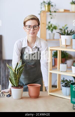 Vertikale Porträt der modernen jungen Frau trägt Schürze stehen von Holztisch mit Topfpflanzen und Erde bereit für die Verpflanzung, Home Gartenarbeit Konzept Stockfoto