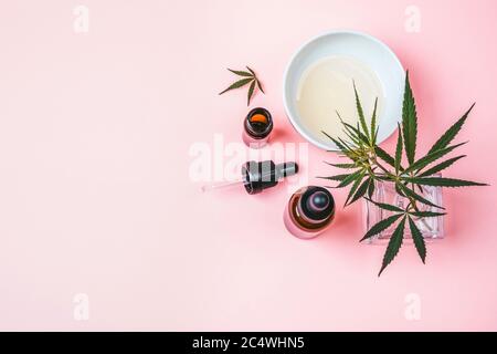 Hanf-Zweig in einer Glasvase, Cannabisölflaschen und eine Tasse mit Öl auf einem rosa Hintergrund Stockfoto