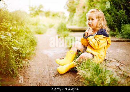 Kleines blondes Mädchen in gelbem Regenmantel stehend und Gummistiefel sitzen auf einer Straße nach einem regen. Sommer, glückliche Kindheit. Stockfoto