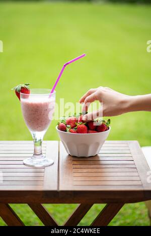 Frau auf einer Sonnenliege, die im Sommer eine Erdbeere aus einer Schüssel neben einem Erdbeer-Smoothie nimmt Stockfoto