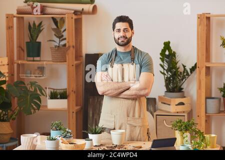 Taille up Porträt von schönen bärtigen Mann trägt Schürze und Blick auf Kamera, während am Tisch mit Zimmerpflanzen und Gartengeräte stehen, kopieren Raum Stockfoto