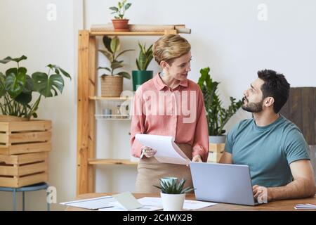 Portrait einer lächelnden Assistentin, die Dokumente einem hübschen bärtigen Mann zeigt, der im modernen Bürobereich am Schreibtisch arbeitet, Kopierraum Stockfoto
