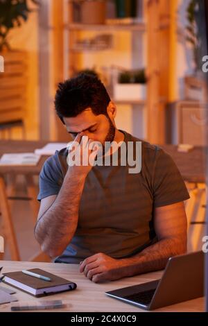 Vertikales Porträt von erschöpften bärtigen Mann, der Augen reibt, während er spät in der Nacht im Home Office arbeitet Stockfoto