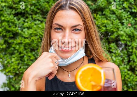 Weibliche Porträt von jungen Erwachsenen trinken farbigen Cocktail an der Außenbar mit Gesichtsmasken - Neue normale Lebensstile Konzept mit glücklichen Menschen toasting dri Stockfoto