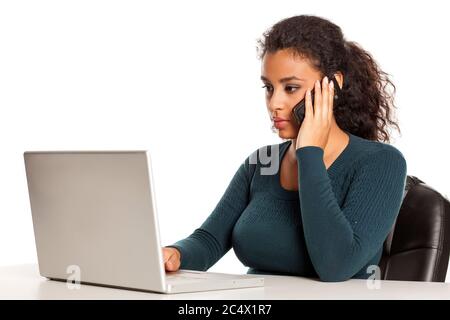 Schöne dunkelhäutige Studentin, die am Telefon spricht und auf ihrem Laptop auf weißem Hintergrund arbeitet Stockfoto