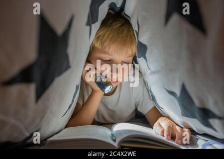 Ein kleiner Junge liest nachts ein Buch mit einer Taschenlampe unter den Deckblättern. Stockfoto