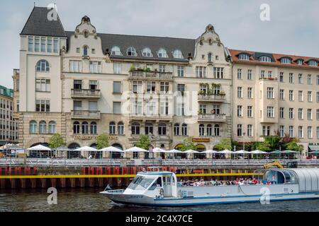 Elegante Gebäude am Fluss mit Restaurants und Bars im Freien auf der Schiffbauerdamm Straße von der anderen Spree und Touristenboot, Berlin, Deutschland. Stockfoto